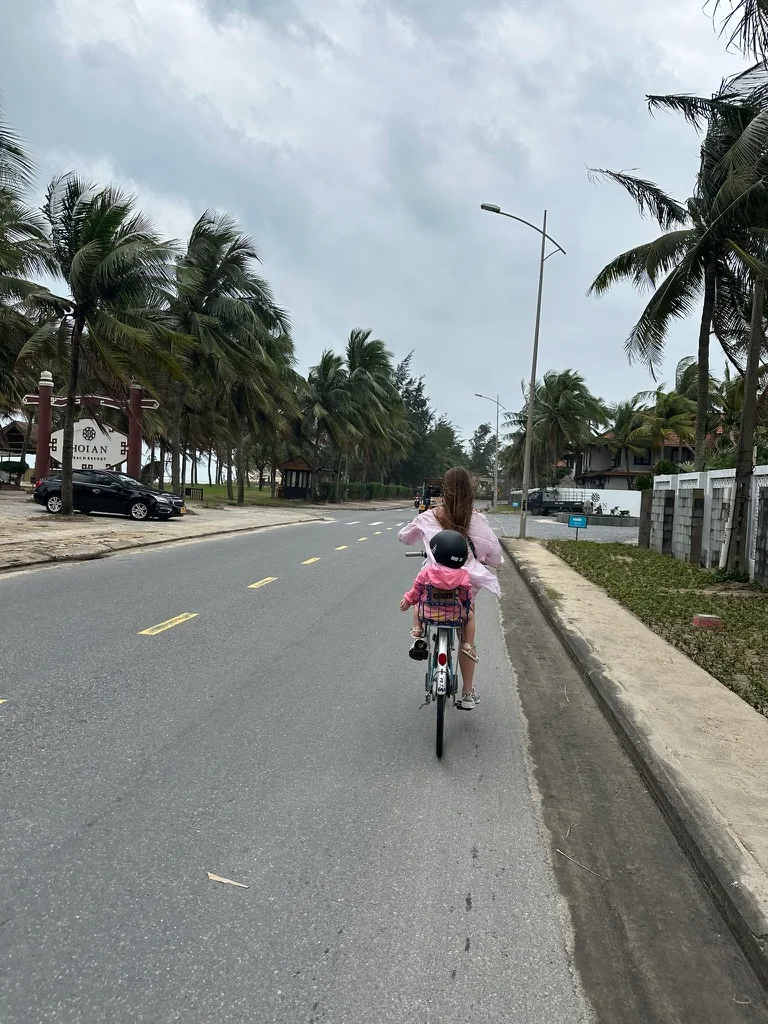 A parent cycling with a child in a rear seat, both seen from behind on a palm-lined street in Hoi An